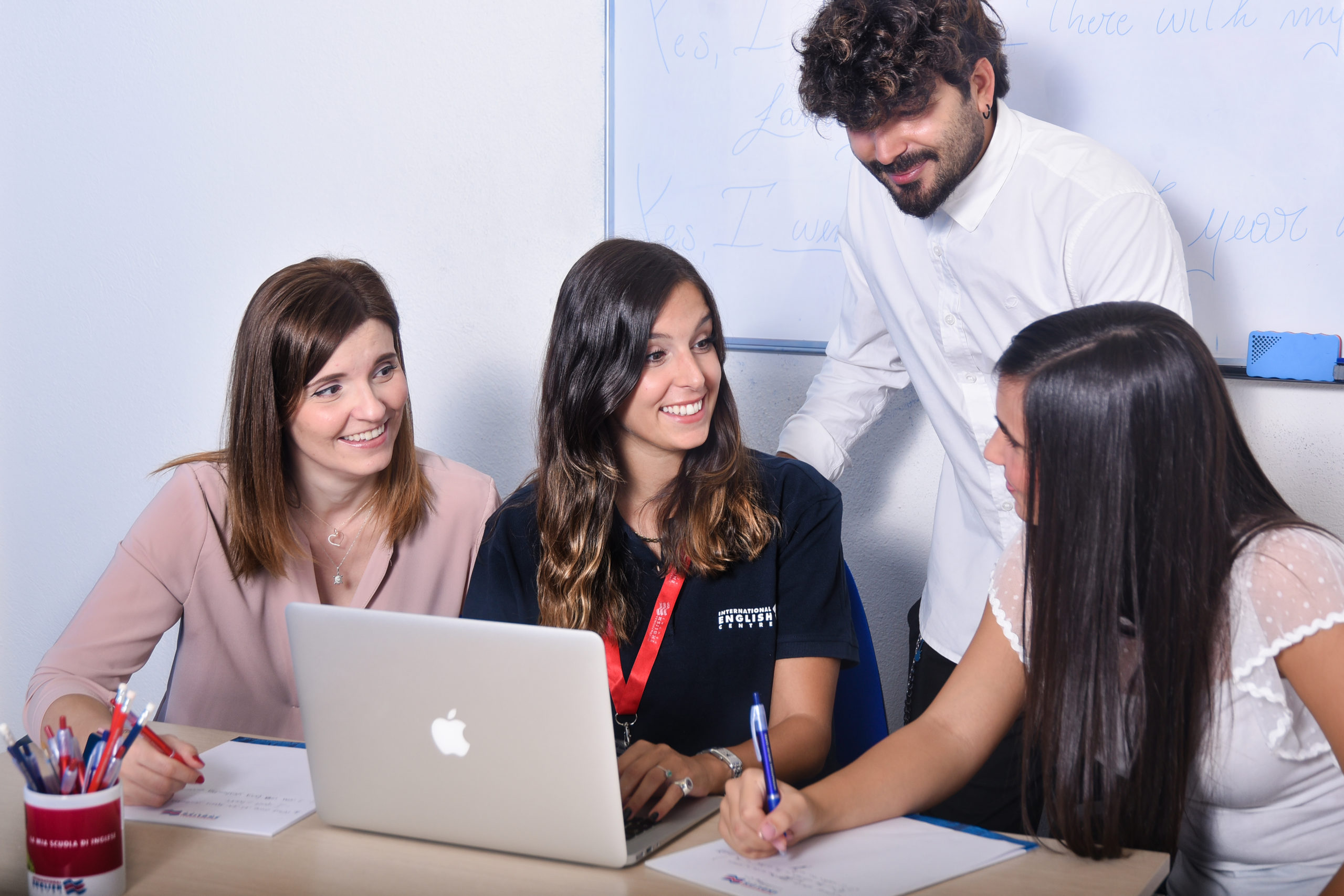 La foto ritrae un docente International English Centre nella sua scrivania durante un corso, circondato da 3 studenti. Nello sfondo la lavagna